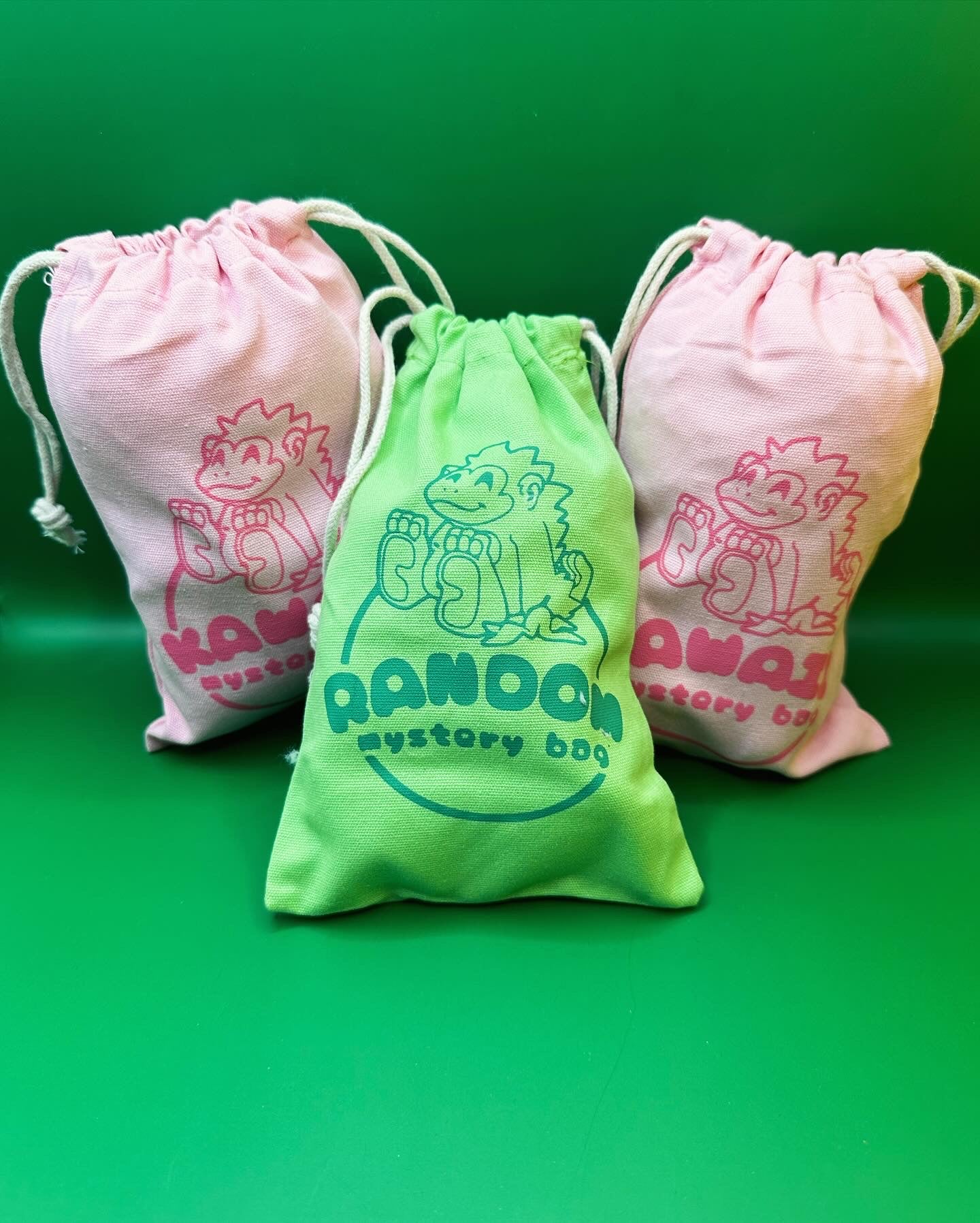 A photo of three mystery bags, one green and two pink. The pink bags are printed with a pink gorilla logo and say “kawaii mystery bag”. The green one is printed with a green pink gorilla logo and says “random mystery bag”