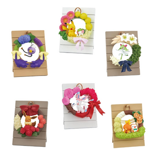 All six types of figures in the wreath collection. 1. Drifloon hanging from a purple and green wreath with bow decoration. 2. Pikachu and Shaymin on a pink and yellow wreath with bow. 3. Kirlia dancing with a green, cream, and white wreath with a navy bow. 4. Eevee relaxing on a red, brown, pink, and white wreath with red bow. 5. Sylveon winking on a heart shaped wreath in pink and red. 6. Torchic and Rowlet napping on a green, white, and brown wreath with orange slice accents.