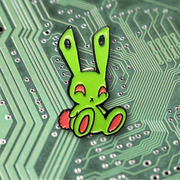 An enamel pin of Bunn-E, the green and pink rabbit with wall plugs for ears! He has a happy expression on his face and he is in a seated position. He's mostly bright green with bright pink fluffy tail, pink feet, and pink eyes. The product is sitting on a background that looks like a faded printed circuit board.